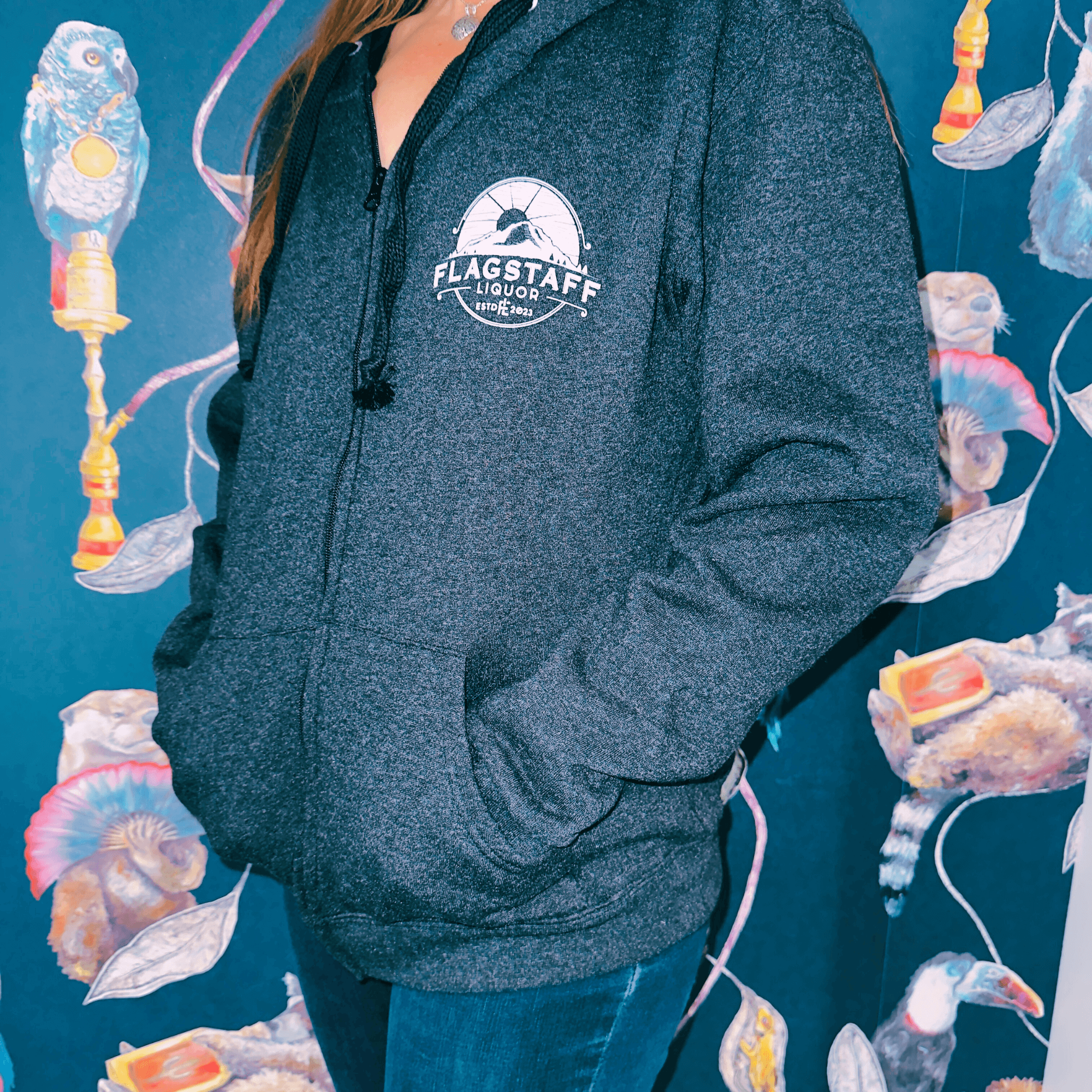 Woman wearing Flagstaff Liquor Mountain Logo Full Zip Hoodie in medium against a colorful mural backdrop, showcasing warmth and style.